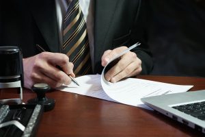 Close up of a man hands as he signs legal document son the desk in front of him.